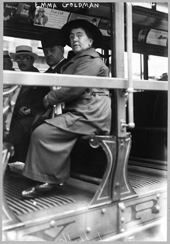 A photograph shows Emma Goldman sitting on a bench in a street car. Two men are sitting next to her on the bench.