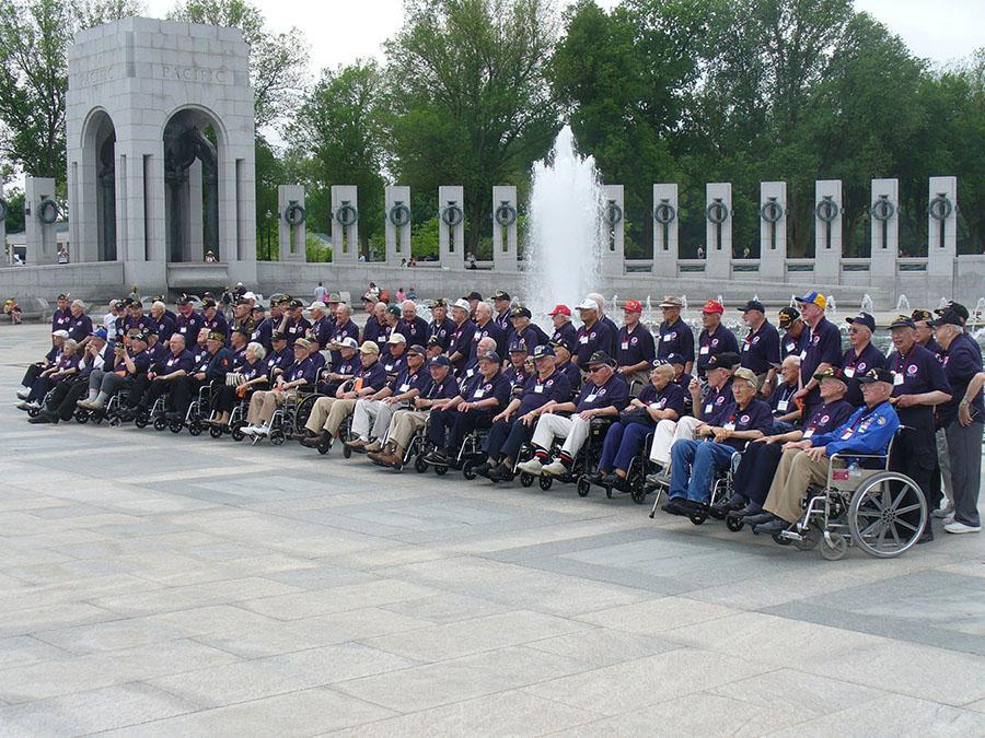 A group of elderly men, many in wheelchairs, all dressed in blue shirts and baseball caps, are shown standing and sitting in a memorial setting, with a fountain and pillars behind them.