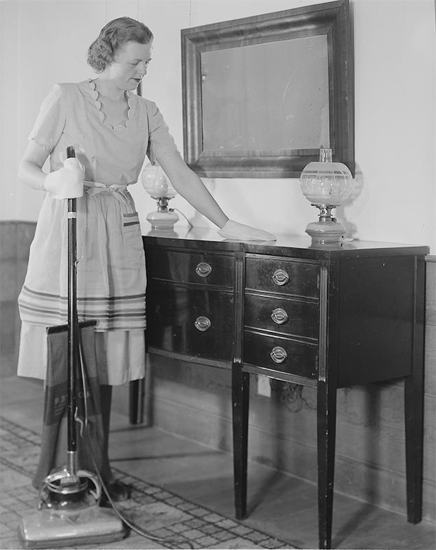 An American woman is dusting a sideboard. She is holding an electric vacuum cleaner in the other hand.