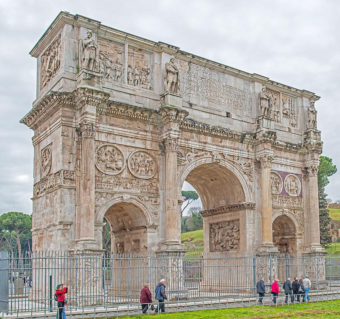 A picture of the face and one side of a large, rectangular marble arch is shown. There are three bays – a large one in the middle and two smaller ones on each of the sides. Above the middle bay there are words carved into the arch. The right and left sides at the top show carvings of figures in various scenes. The one side shown at the left in the picture has carvings at the top as well. Above the two smaller bays there are two circles with figures at various events carved. The one side shown also has a circular carving and a plain wall below the circular carving. Four carved and decorated columns are positioned on the face of the arch – one on each end and one in between each bay. They run from a bottom pedestal to the top where a carving of a person sits at the top. Inside the bays there are carvings of scenes with people as well as sayings. In front of the arch is a gray spiked fence and eight people walk toward the right. They appear to be older and wear pants and coats. Two people stand to the left holding a camera. In the background there are hills, trees, a pale blue sky and other people walking.