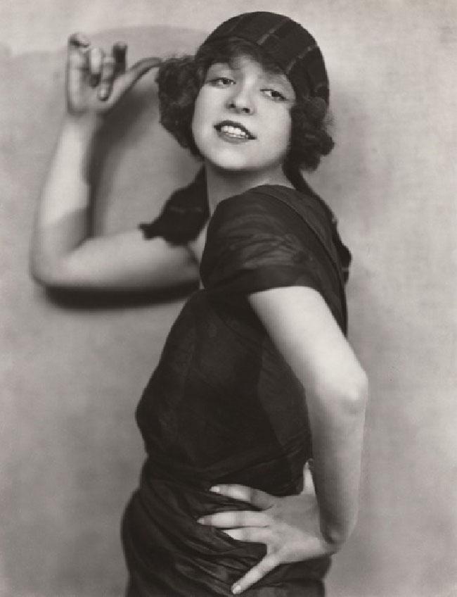 A woman wears a dark dress and striped hat. She smiles and stairs into the distance. Her left hand is on her hip and her right hand is beside her head. She appears to be intentionally posing for the camera.