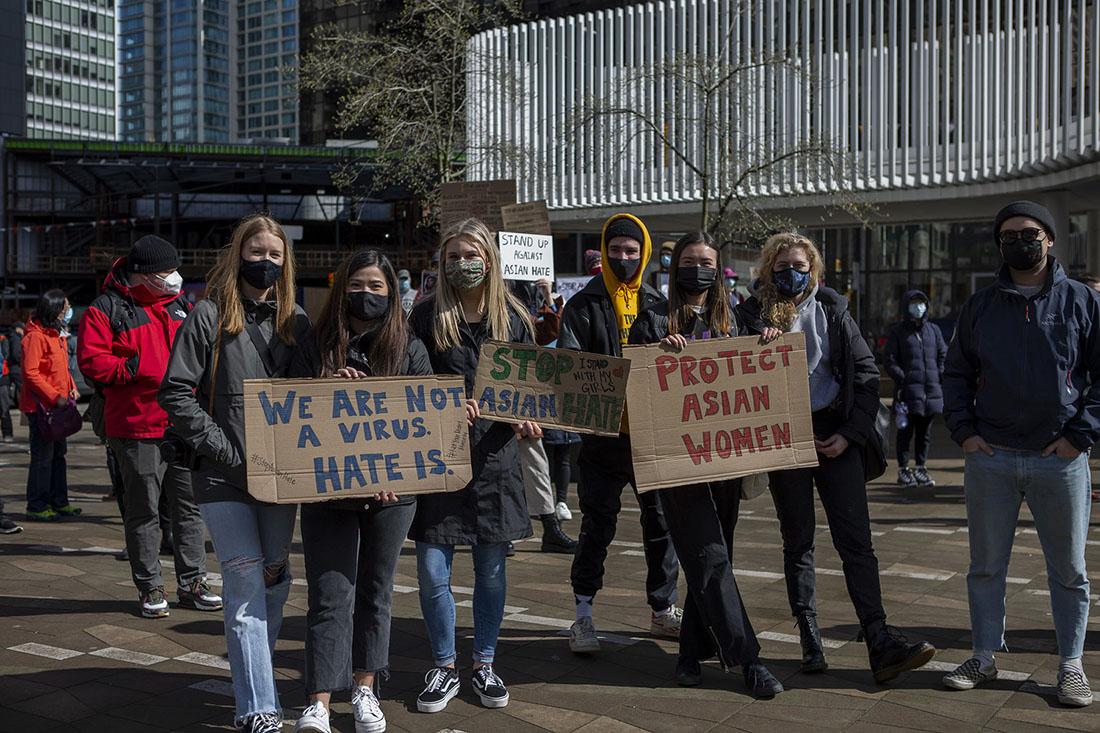 Several people stand wearing masks and holding signs. One sign reads, “We Are Not a Virus, Hate Is.” And another reads “Protect Asian Women.”