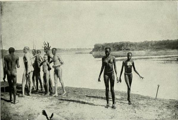 A black and white photo of members of the Nuer people taken in 1906. They stand together on a riverbank, most in a cluster, but one pair off by themselves.