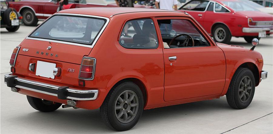 A picture of the side of a small hatchback, two-door, orange car is shown on white pavement. The car has black wheels with gray hubcaps, a black and silver back bumper, a rear-view mirror on the right side of the front hood, and red lights with silver grates on the back. The word “Honda” is across the back in silver lettering. A driver is sitting inside the right side of the car in front of the steering wheel with the window rolled down. Other orange and red cars are seen in the background as well as a man in a white shirt walking.