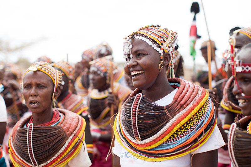 A group of  Rendille women at a festival. All are dressed in traditional, elaborate, colorful clothing with beaded headdresses. Each has a brightly colored striped sash around their shoulders.