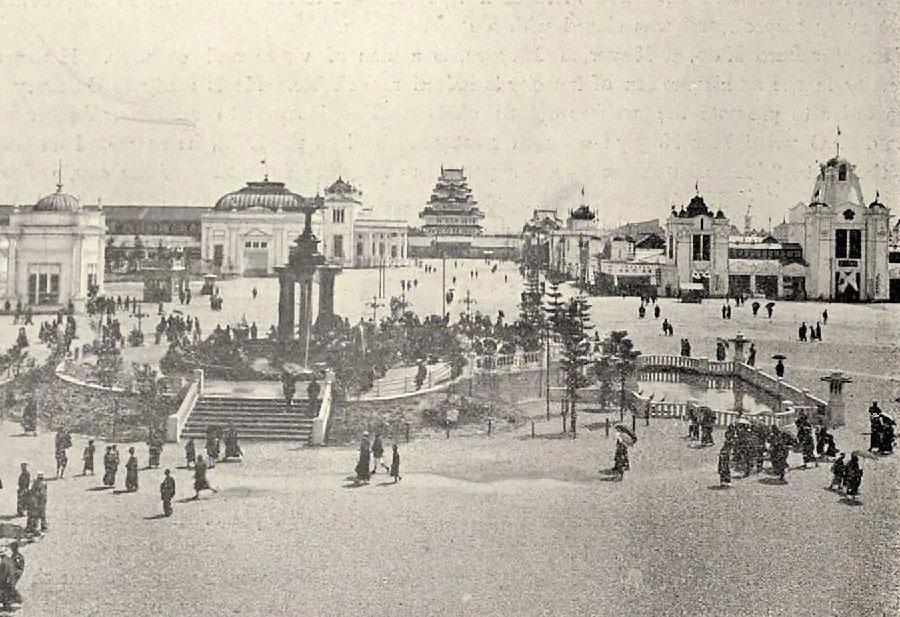 This photograph shows people visiting a public park. A bandstand sits in the center of the photograph. Buildings are visible in the background.