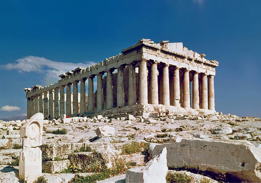 A picture of a rectangular, columned building in ruins is shown against a blue sky with few white clouds. Tall off-white stone columns surround the perimeter of the structure, with broken stone pieces at the top and no roof. Pieces of a stone structure can be seen on the inside. Stone pieces litter the forefront of the image with some green vegetation growing in between.