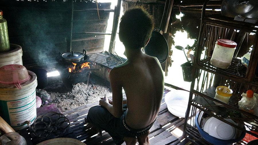 A young person is pictured in a small structure without complete walls or doors. A pot sits over an open flame, and a few bowls and buckets are arrayed on shelves and on the floor.