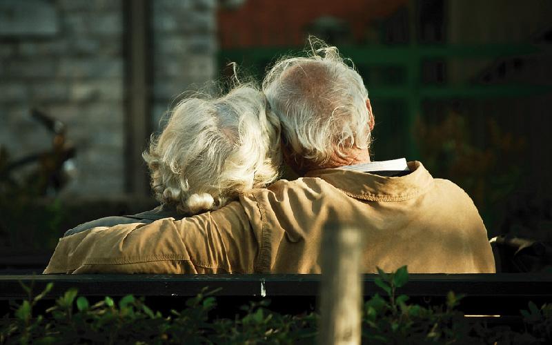 An elderly man and woman are shown from behind sitting on a bench. The man is shown wrapping his arm around the woman’s shoulders.