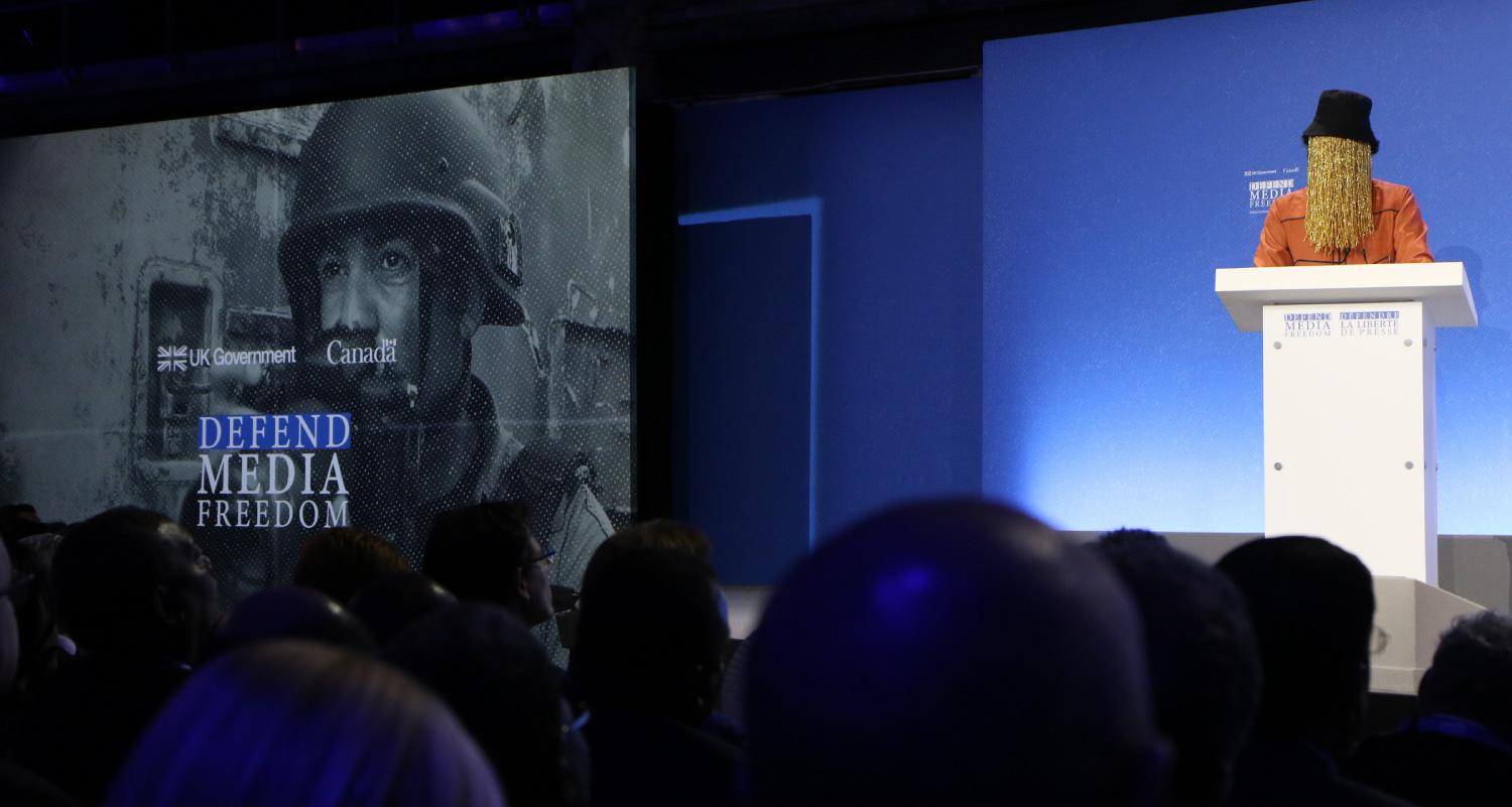 Person standing at podium giving a speech, wearing a tall hat and a veil fully covering their face. An image on a screen to the side of the podium includes the words “Defend Media Freedom”.