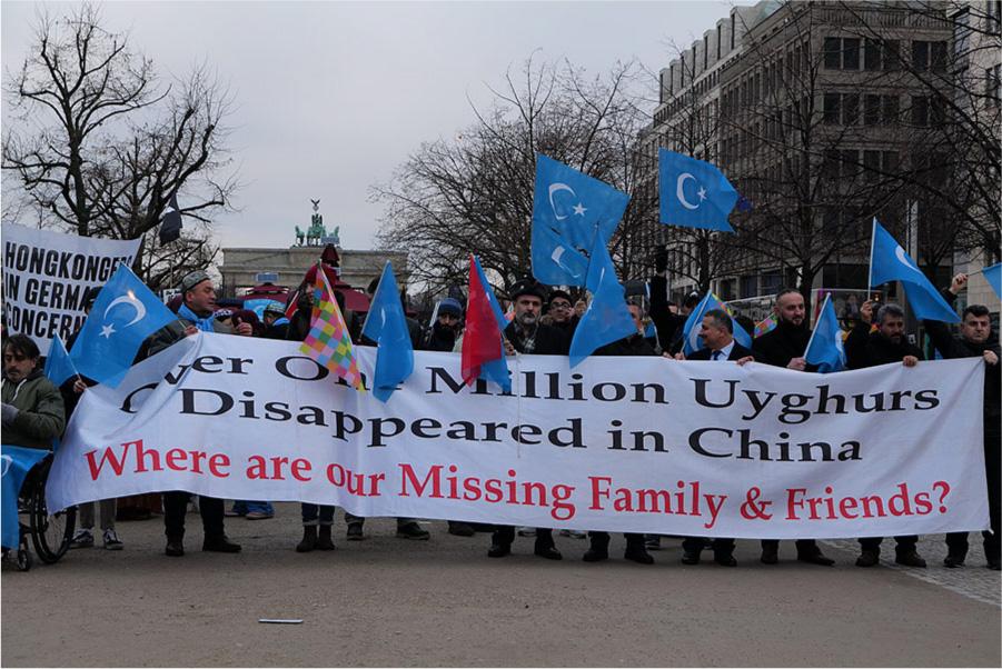 A picture shows people carrying a large white banner on a street in front of a large monument with a green statue on top, trees, and a tall building on the right. The banner reads “Over One Million Uyghurs Disappeared in China” in black and then “Where are out Missing Family & Friends?” in red across the bottom. Several people hold the banner wearing suits, coats, hats and scarves. They also hold flags as they carry the banner. The flags are mostly blue with a moon and star on them, and three are checkered in various pastel colors and one is red. At the left of the banner a man in a wheelchair holds a blue flag. In the left back is a white banner with the words “Hongkingers in Germa Concern” on it, with some of the letters hidden by other flags.