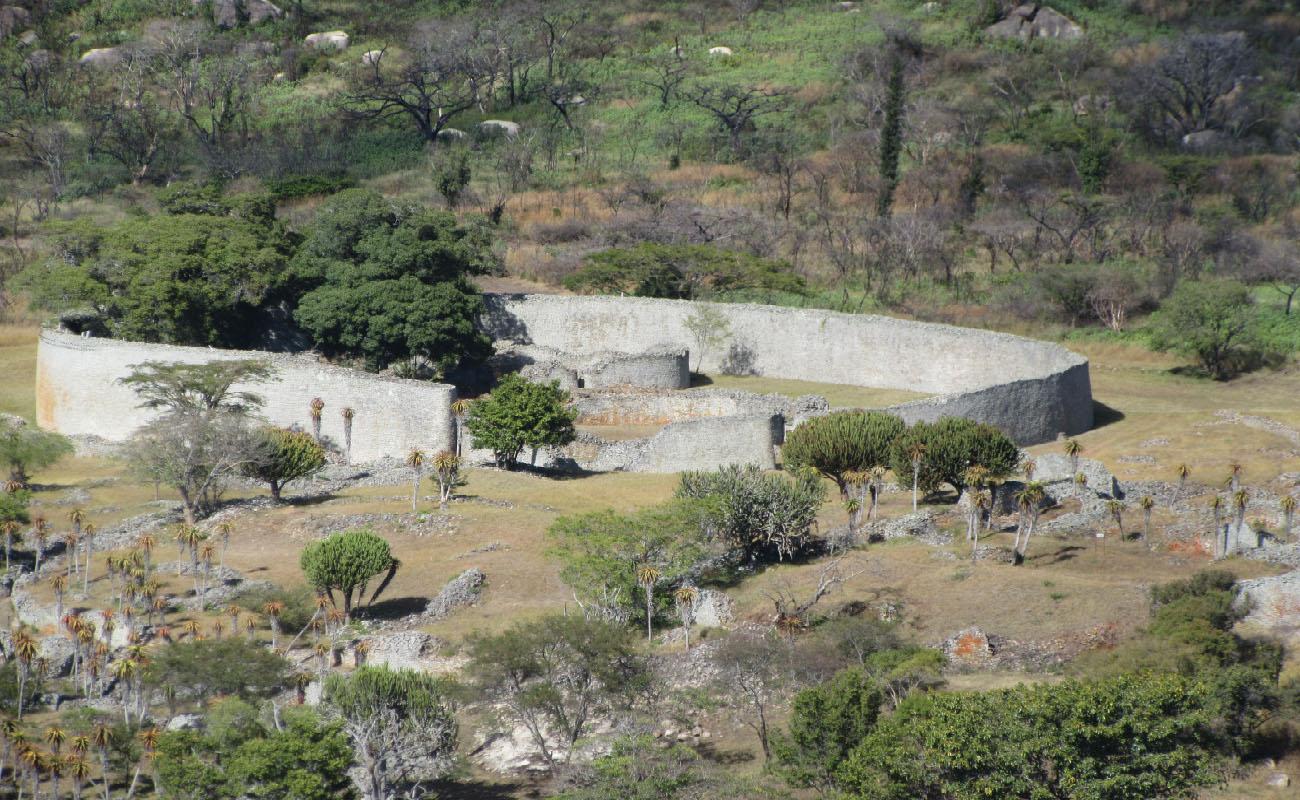 An image of a photograph is shown with trees, grasses, and large white oval stones in the background. A large oval off-white stone wall is shown with a piece missing in the front. Two smaller oval walls are seen inside and large trees grow on the left side. The forefront of the image shows green trees, rocky terrain, and small trees with orange tops.