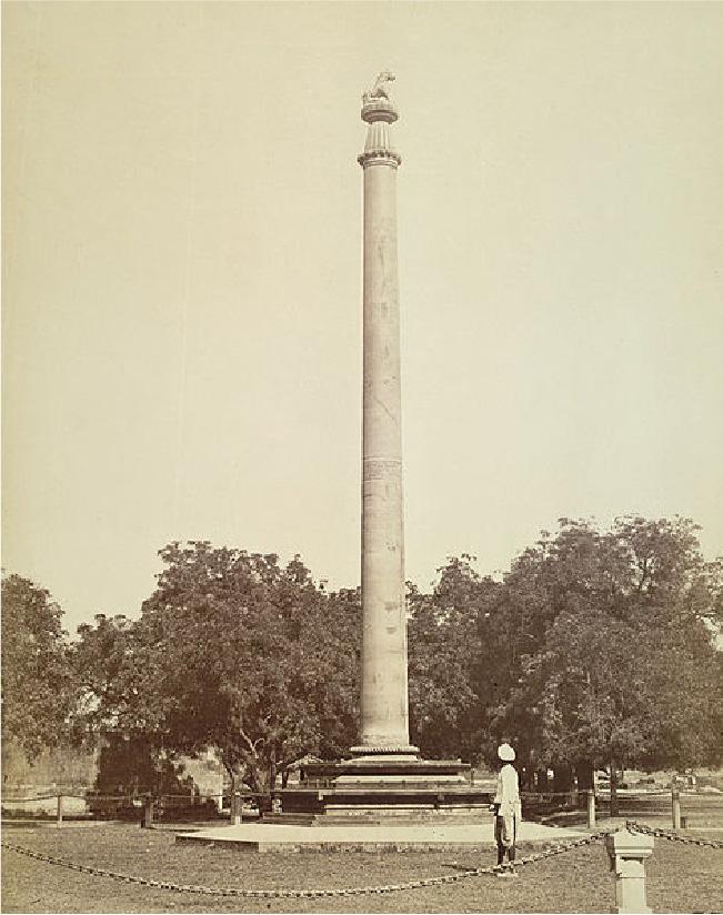 A sepia colored image is shown of a tall, thin spire on a layered, square pedestal. The top of the spire shows a vertical lined conical top, with a round circle above it and a carved object resting at the top. Trees line the background and a chain fence attached to stone pedestals shows in the forefront. A figure in white clothing stands looking at the spire wearing a white hat.