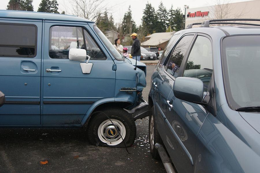 A van and a car are involved in an accident in a parking lot. The van has a smashed front and a flat tire, and glass is on the ground.