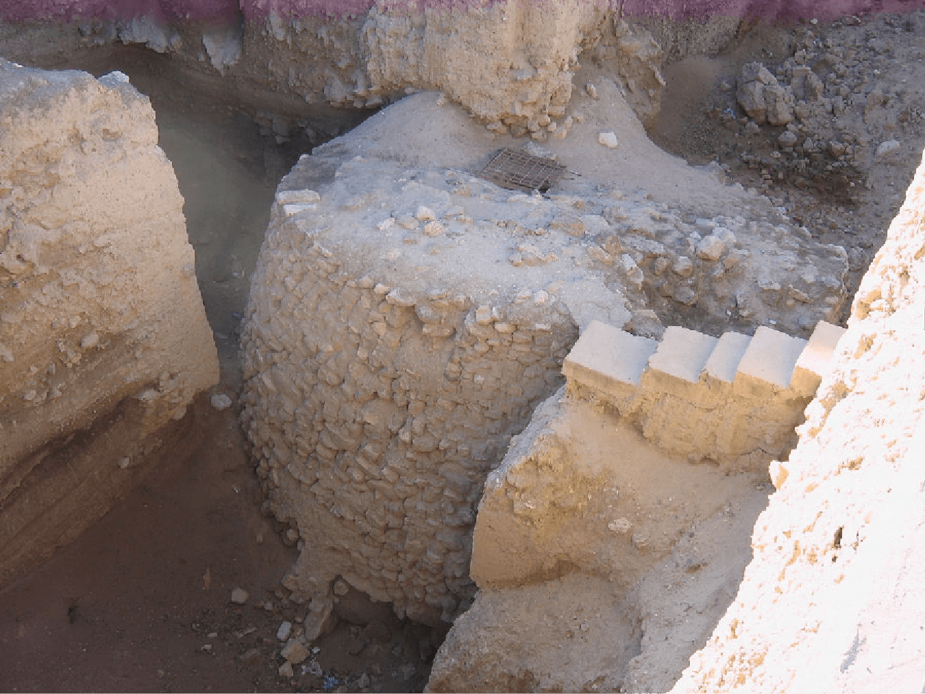 A picture shows an aerial view of a round, off-white stone structure in ruins, with other rocky walls surrounding it. In the bottom middle there are some tiny colorful objects strewn on the ground, five steps are seen in the middle right, a rocky road is seen in the top right, and rocky walls surround the round structure on the top and left. A square grate is seen in the middle of the round structure over a hole.