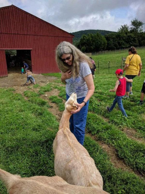 A woman pets a goat, which looks up into her face. In the background, people walk toward a large barn.