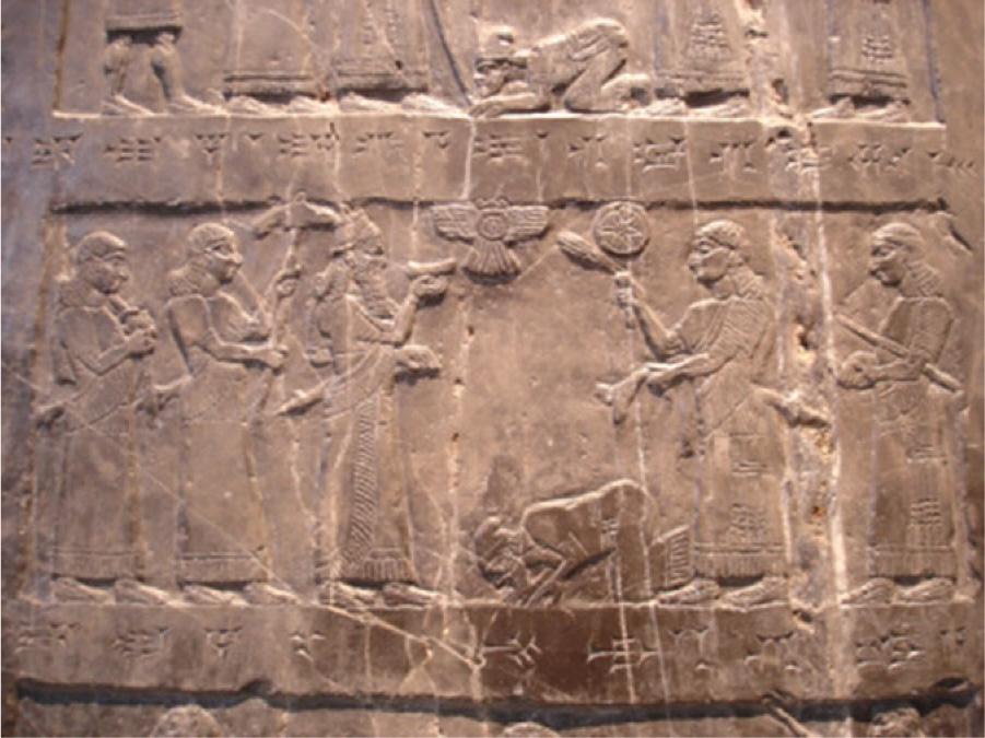 A picture of a brown stained, scratched, and cracked stone is shown. There are two rows of scenes shown. The top row is cut off partially from view, but feet can be seen as well as the bottoms of robes. A person kneeling low to the ground with his head resting on his hands is shown among the feet. In the second row there are five people standing, three facing to the right and two facing to the left. They wear long robes and each carry some type of object. A person is seen in between the two groups kneeling and lowering their head to the ground. Above the kneeling person is a bird shown with spread wings. In between the two rows are symbols etched into the stone.