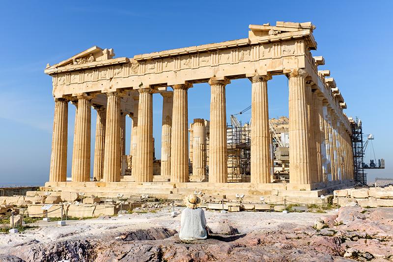 A person sits on the ground in front of the ruins of a large rectangular marble temple with many tall columns supporting what remains of the roof.