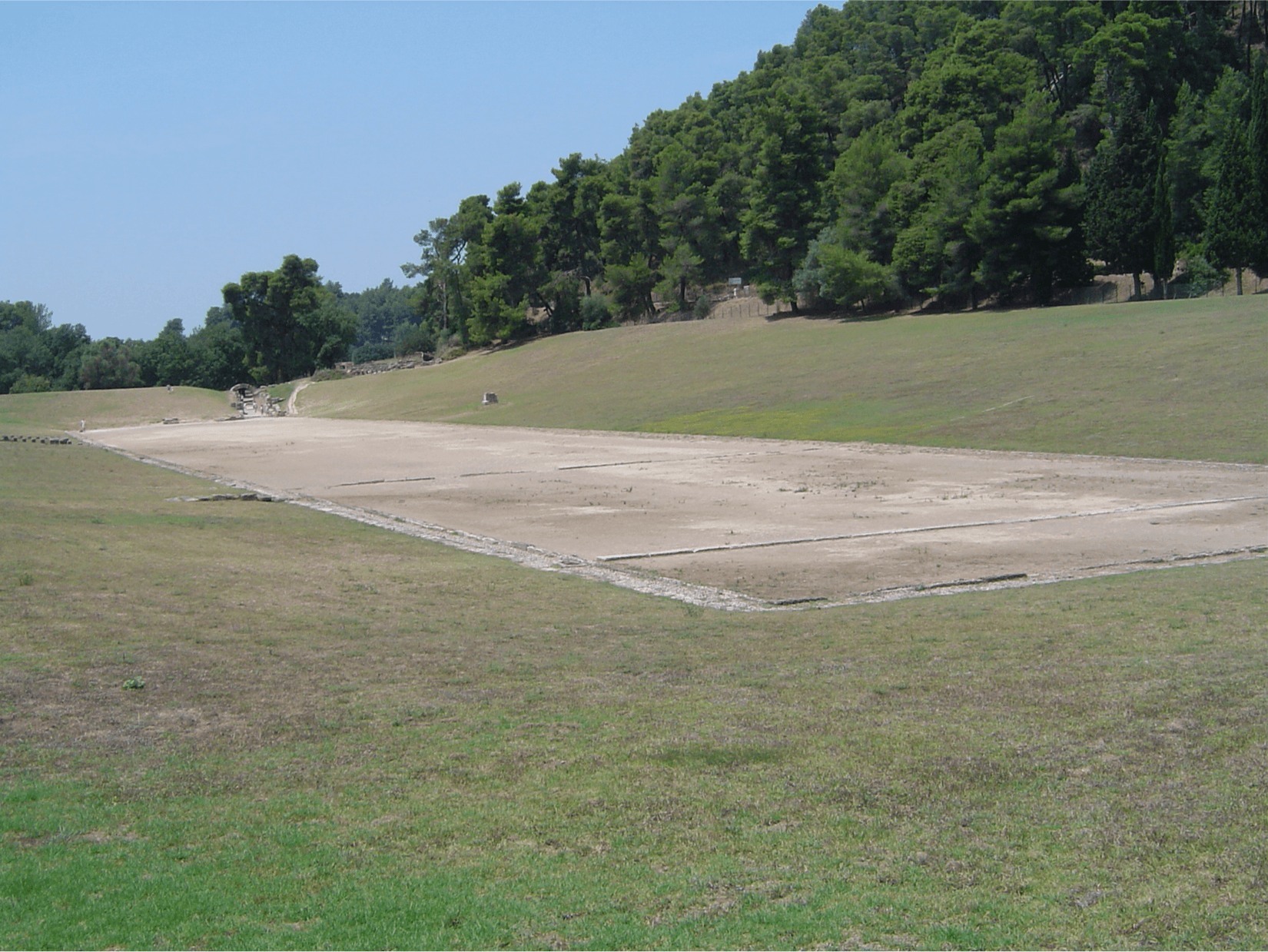 An image of a rectangle section of clay in the middle of a field of green and brown grass is shown. Green trees line the backdrop and a blue sky is seen in the top left section of the image.