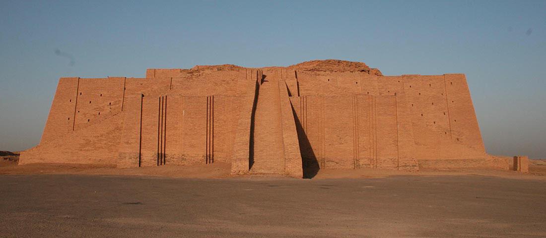 A picture of a very large brown brick structure is shown against a clear blue sky and dark, sandy ground. An immense staircase is shown in the middle front with brick walls on both sides. Lines run vertically in the front of the structure on both sides of the staircase. Square and triangular tiers can be seen on both sides and small rectangular windows are scattered on both sides. Rocky piles can be seen at the middle and right tops of the structure.