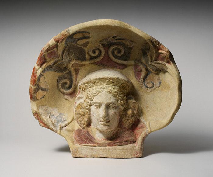 An image of a beige stoneware shell sitting upright is shown with red, black, and orange colors inside, with raised decorations. Inside the middle bottom of the shell is a carving of a head with almond shaped eyes, a large nose, and full lips. Wavy hair is seen at the forehead under a domed hat with etchings on the front. Large earrings depicted as a bunch of grapes show at the sides of the head. A reddish collar is seen around the neck. The background is light blue and the shell casts a shadow to the right.