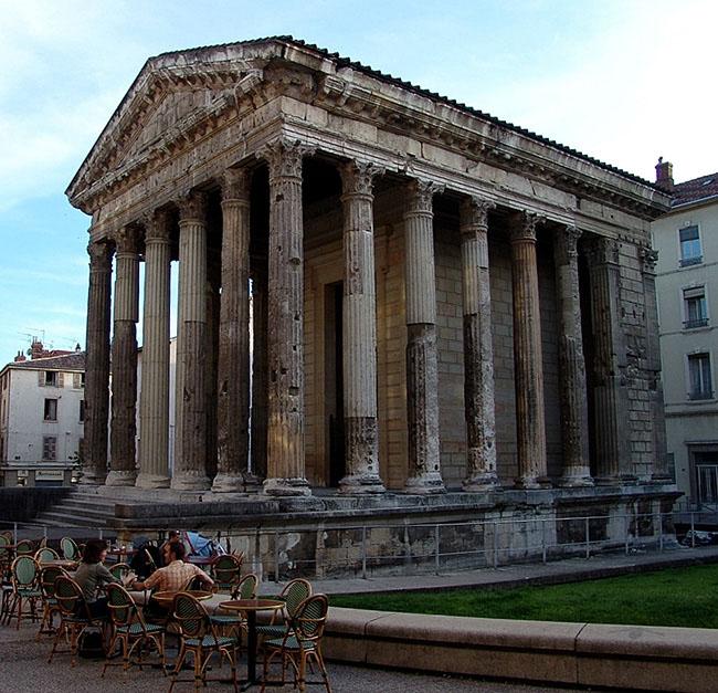 An image of a photograph is shown. A large rectangular building is shown in the middle of the photo with tall columns on the outside perimeter. The columns look worn, faded, and are missing chunks. Inside a stone structure can be seen. The roof is peaked with pointy edges and etchings. It is set on a gray and black stone pedestal with a metal fence surrounding it. Modern-day four-story buildings can be seen behind the structure on both sides with windows and chimneys. Three round brown tables are set in the left forefront of the image with green and brown wicker chairs. A man in an orange and white checkered shirt and a woman in a gray shirt are seated at one of the tables and a person looking into a blue stroller can be seen behind them. Green grass is seen behind a short wall on the right.