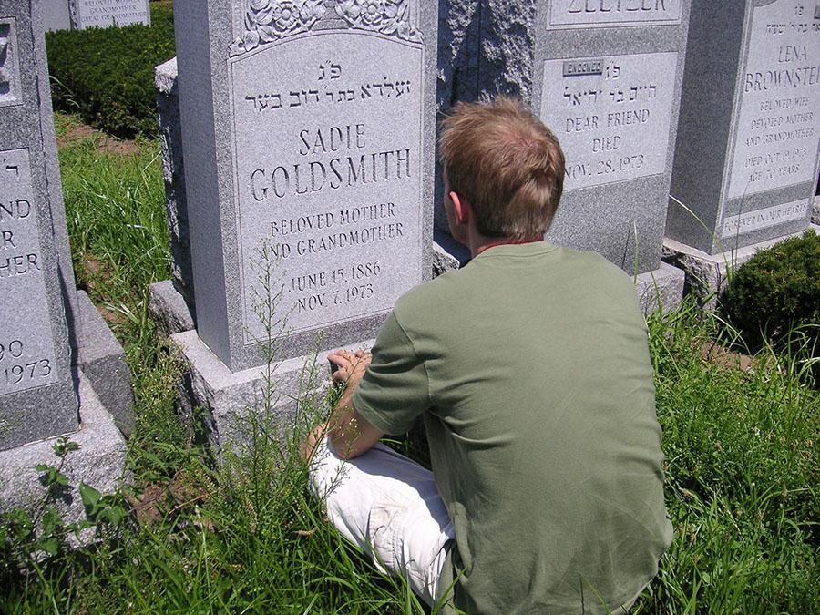 A young man in a green T-shirt and white shorts is shown sitting in the grass in front of a gravestone.