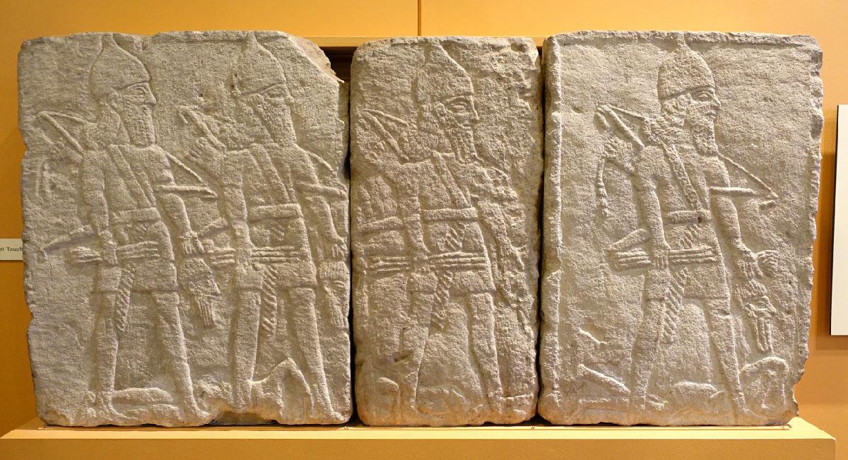 A picture of three rectangular beige stones is shown sitting on a platform with an orange wall in the background. Two figures are in the first stone and one figure is in each of the next two stones. The figures all have long beards, wear rounded, pointed helmets, have sashes across their chests, cloths around their waists, carry bows and arrows, and are facing to the right. Each of the figures hold a severed head with long hair and a long beard in their left hand and bows in their rights. Along the ground lay headless bodies.