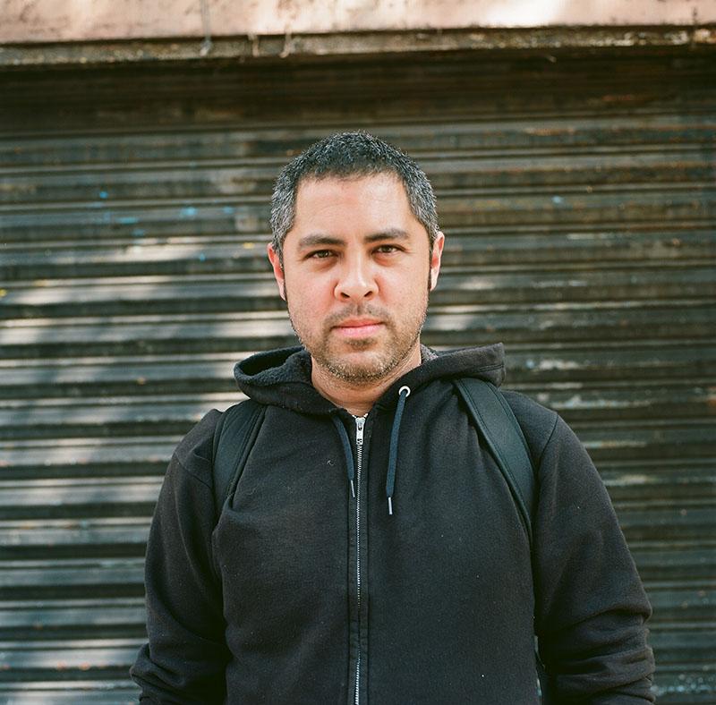 Color photograph of a middle-aged man looking directly into the camera with a relaxed but unsmiling expression. He wears a black, zippered sweatshirt and backpack straps are visible over his shoulders. He stands in front of what seems to be a pull-down metal garage door.