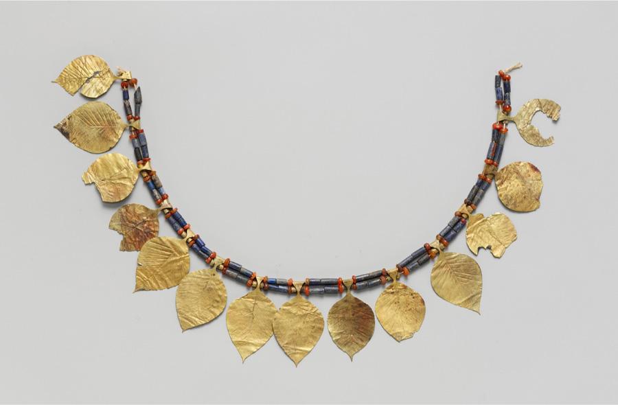 A picture of a U-shaped piece of jewelry is shown decorated with a double strand of black, round, rectangular beads and red circular beads in a pattern. Fourteen gold leaves, some whole and some broken, extend from in between the beads. String can be seen at both ends of the strand in knots.