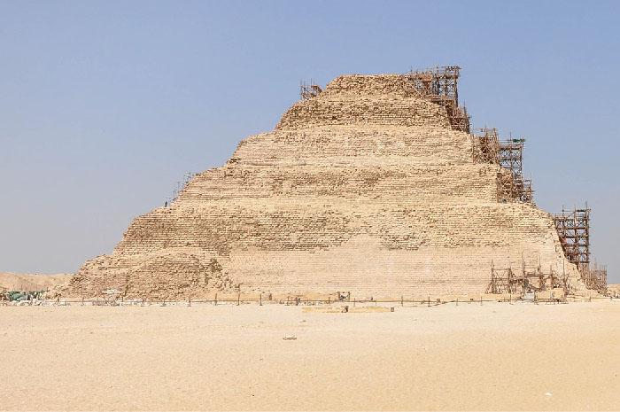 A picture of a brick structure is shown on the background of a blue sky and sandy ground. There are six tiers to the structure, each getting smaller going upward. Scaffolding is shown on the right side of the structure and a simple, short, wooden fence surrounds the structure. No windows or doors are seen.