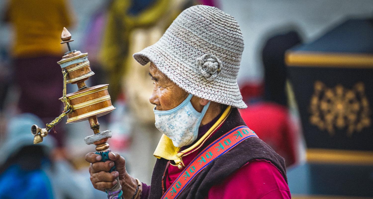 A man wearing a soft low hat and a face mask holds an object with one hand. The object is made up of two drums mounted to a shaft in such a way that allows the drums to rotate, with a wand-type projection mounted to the larger drum.