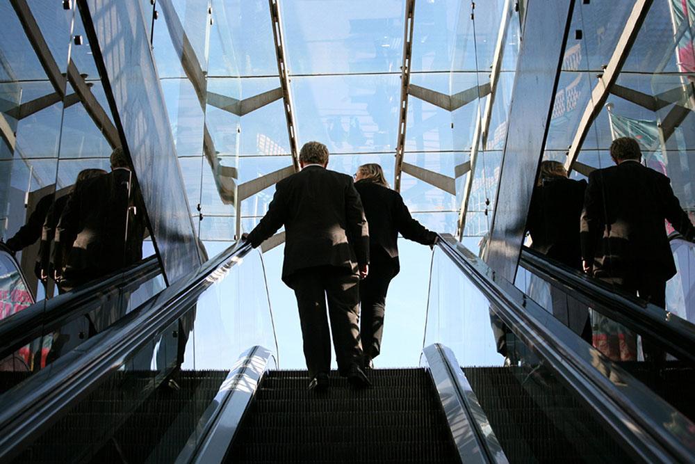 A man and a woman, both wearing business suits, are shown from behind at the top of an escalator