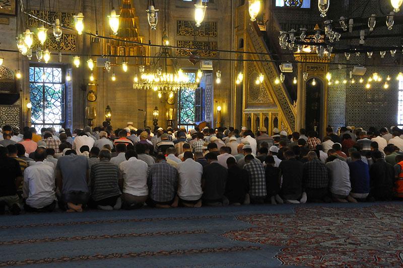 A group of men sit closely together on the floor, all facing the same direction. A circle of lit lanterns hangs from the ceiling.