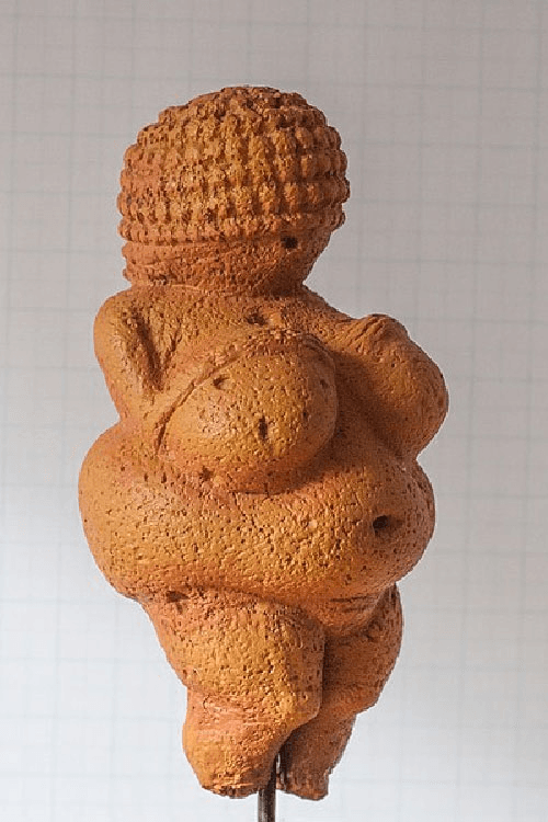 A picture is shown of an orange-colored artifact on a blue and white checkered background. The artifact has a round head, adorned with small squares circling the head. The bottom of the round head shows a small hole. Below the head appear small shoulders, two large breasts, a large protruding belly in the front and back, a deep belly button, and thick legs that turn into toes. A rod is shown holding up the artifact.