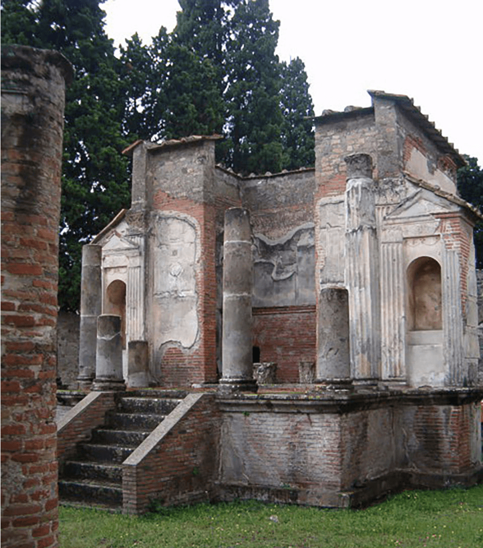 An image of a photograph is shown. In the photo, an old building partially ruined is shown. At the left forefront corner, a tall, round red brick structure is shown, with flaked bricks and black smudges at the top. The ground in the forefront is green grass and a line of very tall trees is shown in the background on a pale sky. In the middle a building stands made of faded, flaked red and white bricks. The building sits on a raised brick platform with black steps at the front. A short red brick wall shows on either side of the steps and broken columns are stationed at the top on either side. Two more columns are shown, one on each corner of the raised platform. Behind the columns a square wall stands with the front missing. Smaller rooms are attached on each side with a rounded arched opening. The walls are gray, black, red, and white. No roof is visible, but the edges of black scalloped tiles can be seen at the tops of the remaining walls.