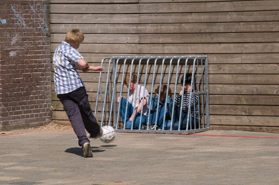 Boy kicking a soccer ball on a playground toward three other boys who are caged against a wall by a small metal goal post. The boys are crying or holding their ears.