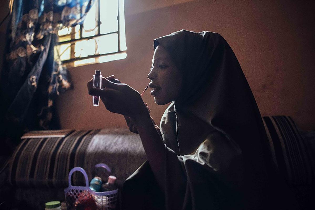 A young woman applies makeup while sitting on a couch. She is wearing a head covering.