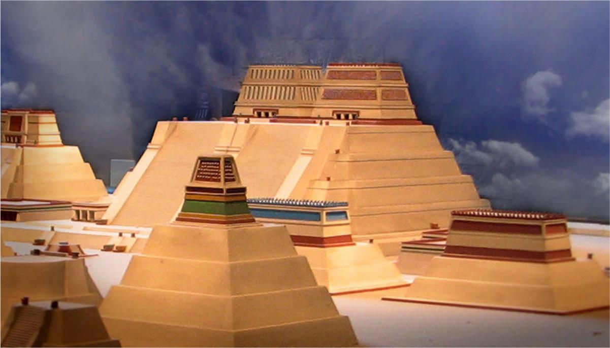 A photo of varying sized building models is shown on a flat white surface and darker blue cloudy background. One large tiered structure shows in the middle back with orange colored lower tiers. Two separate tiered buildings sit atop the structure and show doors on the lowest tier. The left one shows vertical dark lines while the one on the right shows red coloring across the walls. In the forefront shorter and smaller more tiered structures are shown. All have lower orange colored plain tiers with tops tiers decorated with blue and red colors.