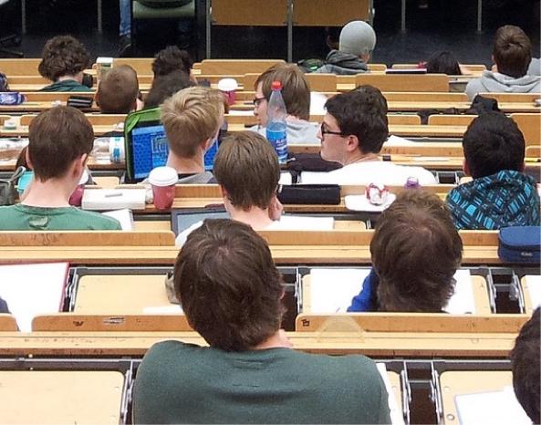 Many rows of students are in a classroom. One student has an open laptop on his desk.