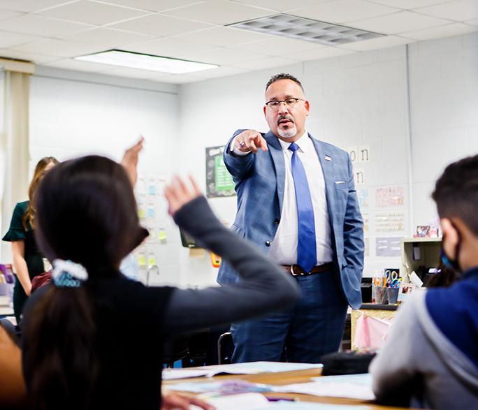 An elementary classroom. A man wearing a suit and tie stands in the center of the room. Students in the foreground hold their hands in the air, waiting to the be called on.