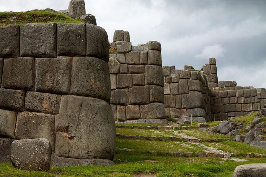 A photo is shown of stone walls standing on green grassy lands with a cloudy blue sky in the background. There are two walls that stand alone while the wall in the background forms a corner and extends out to the end of the photo at the right. The wall stones are square and rectangular. The ends are rounder and the first wall in the forefront has grass growing on the top.