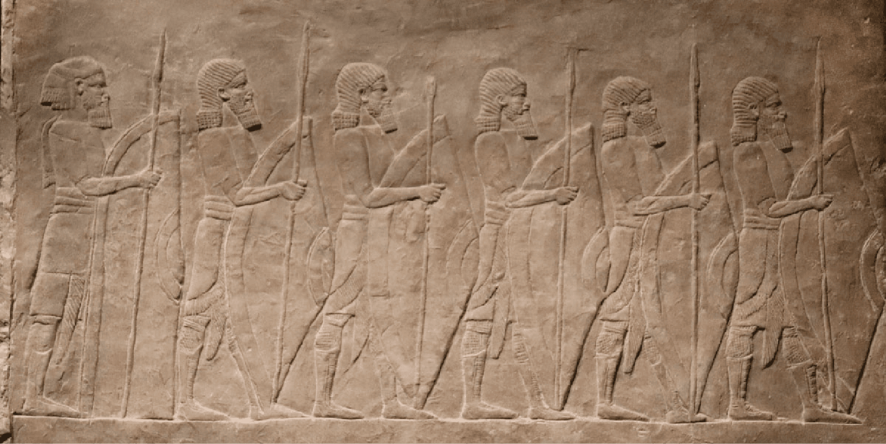 A picture of a stone carving is shown. Six figures are shown facing to the right, all wearing striped headdresses, having long beards, cloths around their waists, and tall boots. They all hold spears in their right hands and full length shields in their left hands.