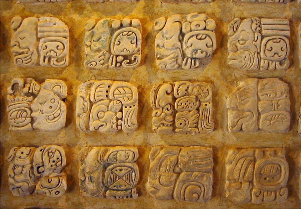 A photo of an orange stone is shown with twelve raised and rounded squares, four across and three down. The flat part of the stone is swirled orange colors. The raised portions are paler orange colored and decorated with various images. Some faces can be seen with large noses, open and closed eyes, beards and moustaches, and large mouths on the squares. Other designs include swirls, circles, squares, notches, and lines.