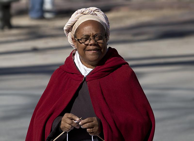 Woman dressed in traditional clothing in Clonial Williamsburg Whe is wearing a  head wrap and cape. She is holding knitting needles.