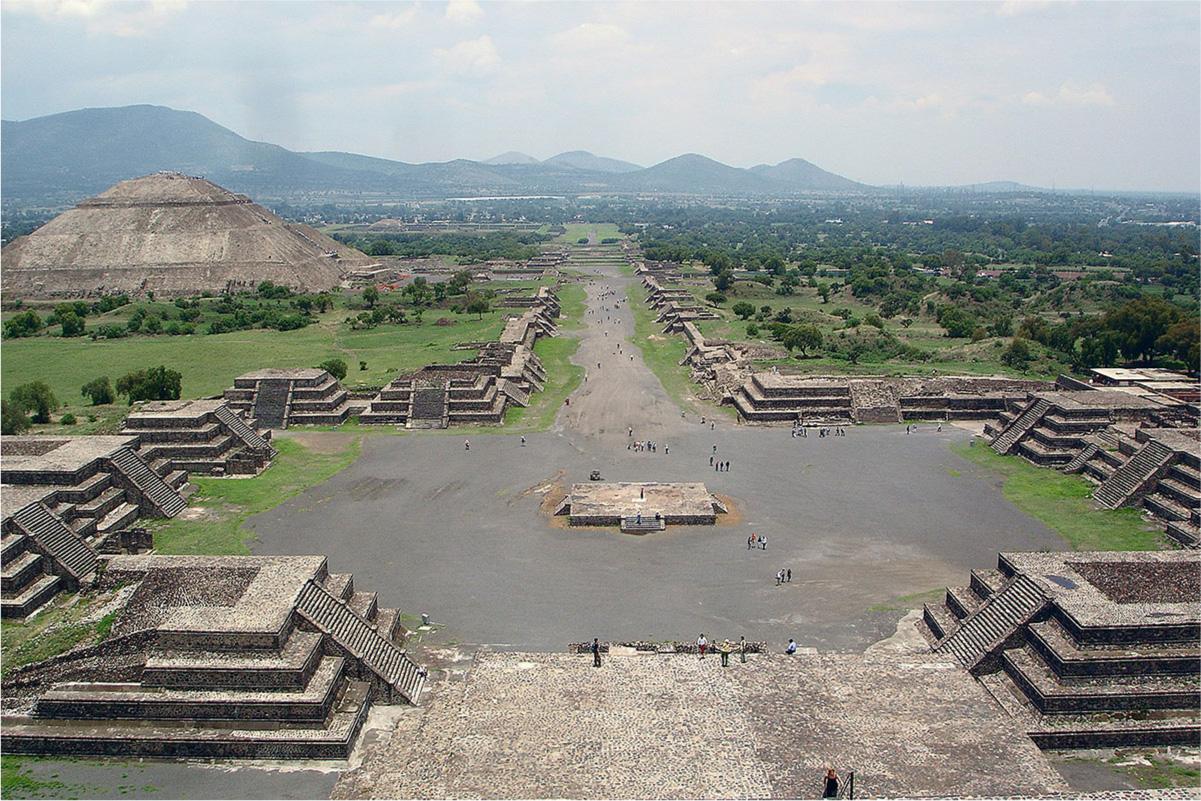 An aerial photo is shown of stone ruins of a city. In the front the ground shows a flat, square, stone area. To either side a four-tiered grayish brown structure stands with large stairs on one side leading to the top, which is flat. Next are three more identical structures set back on both sides as well with a large open dark area of ground in between. A square flat pedestal sits in the middle with a set of stairs facing the front. Next on both sides are two, tiered structures, facing forward, set in closer together with a dirt road beginning between them. As the road goes off into the distance, it is lined with the more tiered structures, in single rows, on both sides. Behind these single structures on the right are trees and other building in in the distance. On the left behind the structures are some trees and then a very large pyramid shaped stone structure behind the trees. The rounded, stone top flattens out and no windows or doors are visible. People are scattered in the open area and walking down the dirt road. The far background shows trees, a city, and mountains.