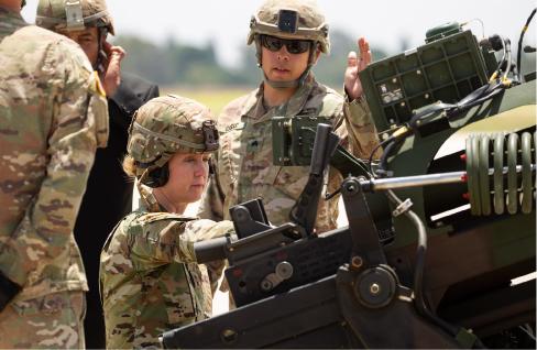 A photograph shows a female soldier and male soldiers standing beside a large gun.