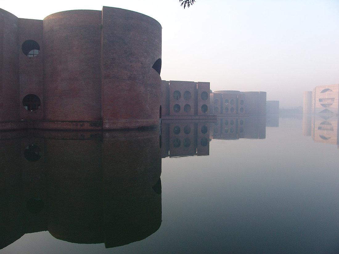 The National Assembly Building portion of the Capitol Complex in Dhaka, Bangladesh. A brick building with rounded walls and large circular open windows sits in a lake. In the background, another building in the complex with an entirely different design sits within the lake.