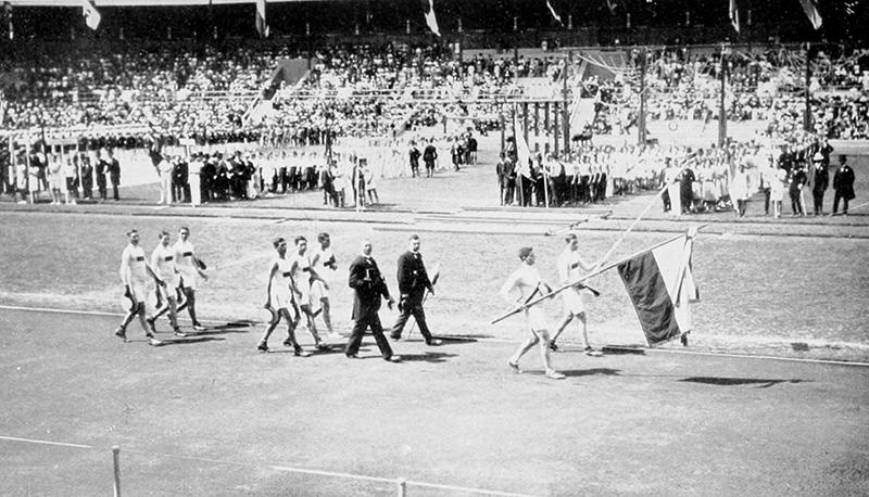 A delegation of people march with a flag. Behind them, other groups of people stand with flags.