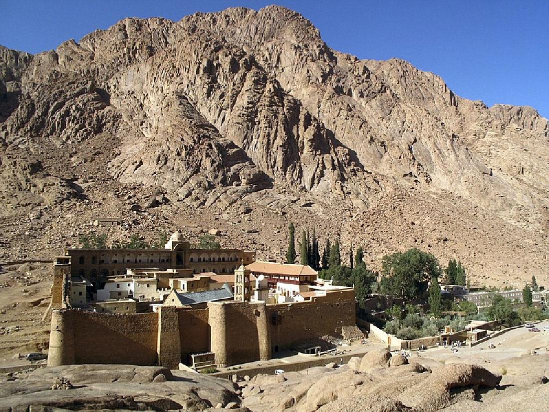 A picture of various buildings built tightly next to each other within square-shaped high brick walls is shown along the backdrop of a tall, brown, rocky mountain with a blue sky in the background. The buildings are various shapes, sizes, and colors, with one of the buildings running the length of the area in the back and taller than the rest. The buildings have windows throughout and are various shades of brown, black, and white. The brick wall around the buildings is brown and has two rounded and one square column in front, one rounded one on the left front corner, and a square tower on the left side rising higher than the wall. Between two of the columns on the front there is a cemented walkway coming out and running the length of the enclosure toward the right of the picture into an area with green trees of various sizes and shapes. Some of the trees are within smaller stone walls, and some rise higher behind the buildings. More short, windowed structures are seen in the far right of the picture. The terrain in the forefront of the picture is rocky and barren. One small building sits on the mountain in the background behind the walled enclosure.